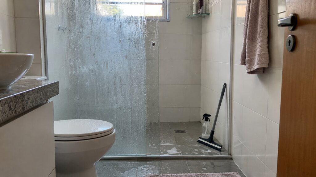 Box de vidro de banheiro com metade manchada e metade limpa, rodo e borrifador no chão